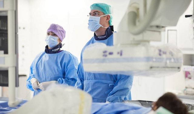 Zwei Ärzte in Operationskleidung in einem Operationssaal der Kardiologie. Auf einem Tisch vor ihnen liegt eine Patientin.