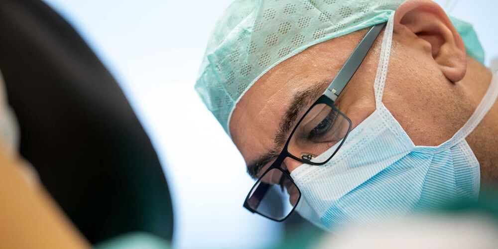 Ein Arzt mit Operationskleidung und Brille schaut konzentriert nach unten. 