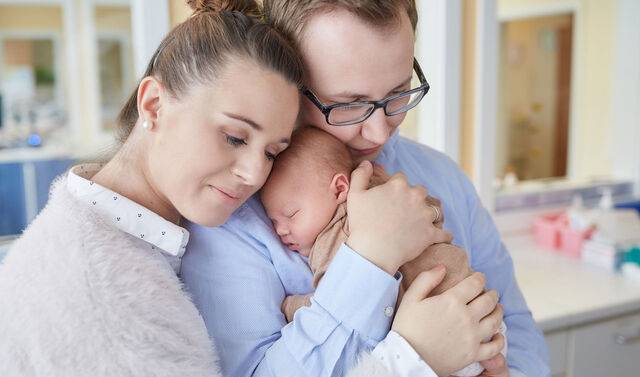 Ein junges Paar das sich umarmt und ihr neugeborenes Baby auf dem Arm hält.