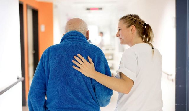 Pflegerin begleitet Senioren durch den Flur und hat ihm eine Hand auf den Rücken.